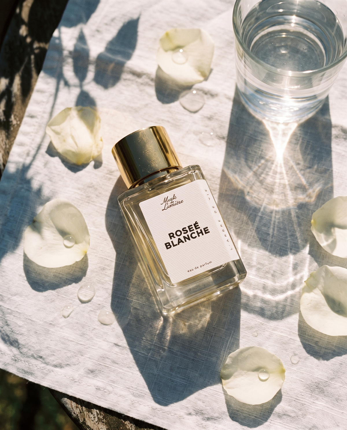 Roseé Blanche eau de parfum by Musk de Lumière on white linen with rose petals and the caustics of a glass of water